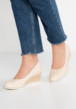 Fødder iført lyse beige kilehælssko kombineret med blå jeans med korte, frynsete ben på en hvid gulv.