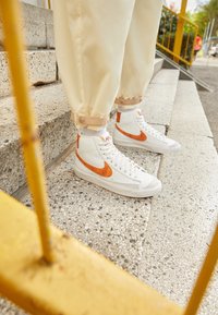 Vita högtoppsneakers med orange detaljer, texturerat läder och meshpaneler. Bärs med gräddfärgade byxor med uppvikta ben på stensteg.