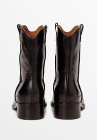 Botas de tobillo de cuero negro con una textura suave, con un borde superior en forma de ondas y un tacón bajo en bloque. Cintas traseras para facilitar su colocación.