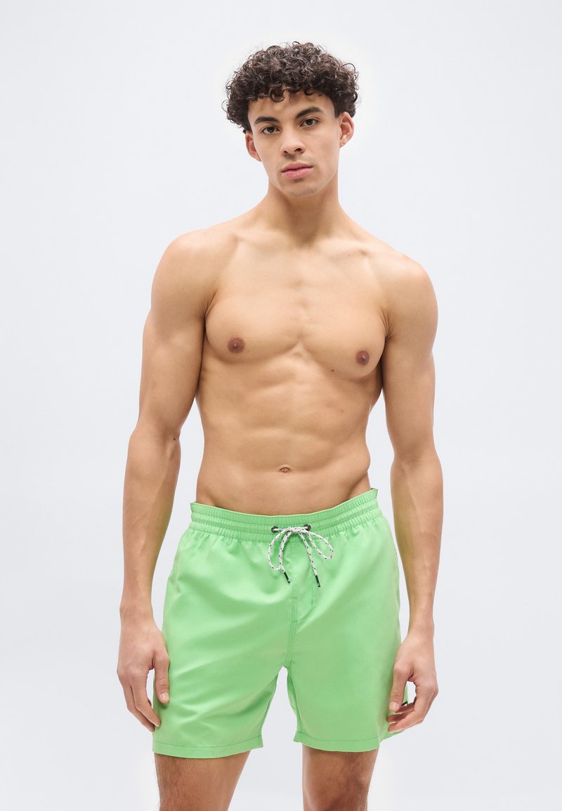 Νέος άνδρας με σγουρά μαλλιά που φοράει πράσινα μαγιό με κορδόνι, στέκεται μπροστά σε απλό λευκό φόντο.