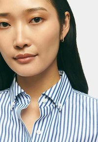Camicia button-up con righe verticali blu e bianche, tessuto morbido e colletto classico. Il modello indossa orecchini a cerchio piccoli.