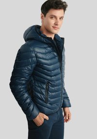 Mężczyzna w granatowej, pikowanej kurtce puchowej z kapturem, zapinającej się częściowo na zamek, z rękami w kieszeniach, stojący na jednolitym tle.