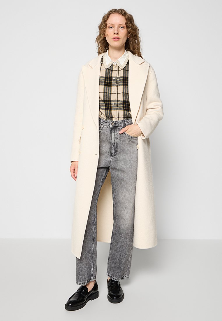 Kremowy płaszcz z klapami, nałożony na koszulę w kratkę w odcieniach beżu, czerni i zieleni, w zestawieniu z szarymi dżinsami i czarnymi mokasynami.