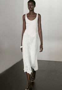 Weißes ärmelloses Kleid mit einem glatten Oberteil und einem strukturierten Spitzenboden. Es hat eine fließende Silhouette und eine Midi-Länge. Schwarze Sandalen.