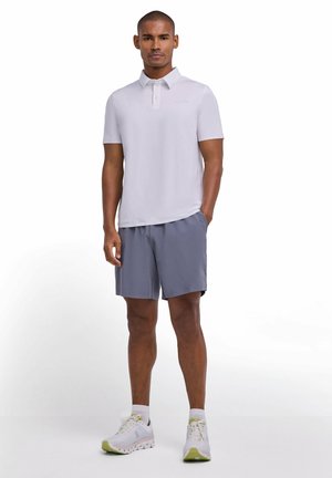 Mann trägt hellgraues Poloshirt, graue Shorts, weiße Socken und weiße Laufschuhe und steht mit einer Hand in der Tasche.