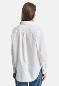 White button-up shirt with a relaxed fit, pleats at the back, and long sleeves. Made from a smooth cotton fabric.