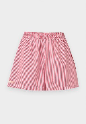 Vrouw met donkere zonnebril, rode top met ritssluiting en getailleerde taille, gestreepte shorts, witte sokken en witte sneakers, staand met één hand op de heup.