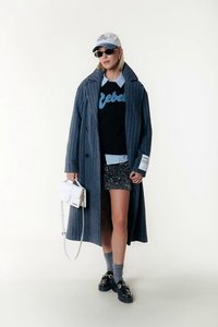 Blue pinstriped long coat, black sweater with “Rebel” print, sequined shorts, gray socks, black loafers, white handbag, and a baseball cap.