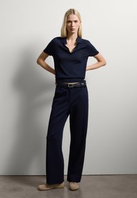 Polo shirt blu navy con colletto e tre bottoni, abbinato a pantaloni blu navy larghi. Una cintura nera con fibbia argentata e scarpe beige completano l'outfit.
