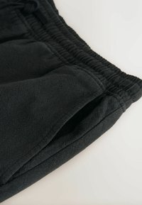 Gros plan sur un pantalon de jogging noir en tissu montrant une taille élastique et une poche latérale sur un fond clair.