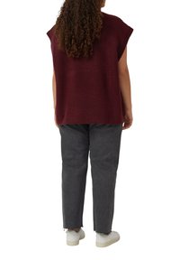 Person mit lockigem Haar, der einen ärmellosen burgunderfarbenen Pullover, dunkelgraue fransige Jeans und weiße Sneakers trägt, steht mit dem Rücken zur Kamera.