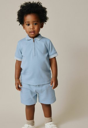 Svetlo modra teksturirana polo majica z zadrgo in belimi poudarki, kombinirana z ujemajočimi kratkimi hlačami. Čevlji so beli z ježki.