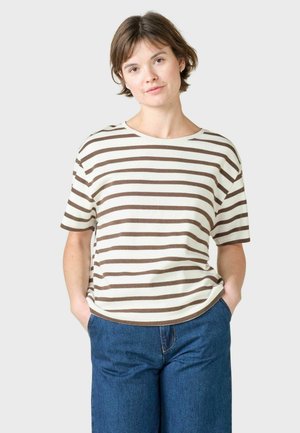 Kvinde med kort brunt hår iført en hvid og brunstribet t-shirt og blå jeans, stående med hænderne i lommerne mod en lys baggrund.