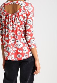 Rode bloemenblouse met een geplooide achterkant en een kleine sleutelopening, voorzien van zwart-witte bloemen op zachte stof. Driekwartmouwen zijn inbegrepen.