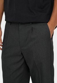 Grau maßgeschneiderte Hose mit strukturierter Oberfläche, ausgestattet mit zwei Seitentaschen und einer klassischen Passform. Kombiniert mit einem schwarzen Hemd.
