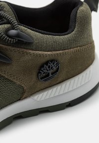 Zielony tekstylny i zamszowy sneaker z czarnymi akcentami, z okrągłym logo oraz białą i zieloną gumową podeszwą z teksturowanymi detalami.