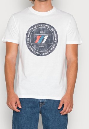 Homme portant un t-shirt blanc avec un logo noir rond contenant des bandes rouges, bleues et blanches ainsi qu'un texte blanc autour.