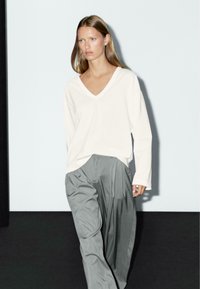 Witte lange mouwen V-hals top in combinatie met losse, geplooide lichtgrijze broeken. Zacht materiaal en relaxte silhouet, met minimale details.