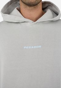 Lys grå hoodie lavet af blødt stof, med en kængurulomme og et hvidt "PEGADOR" logo på brystet. Klassisk design med hætte.