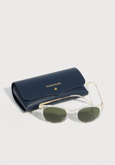 Διαφανή γυαλιά ηλίου με σκουρόχρωμους φακούς δίπλα σε μια σκούρο μπλε θήκη Polo Ralph Lauren με χρυσά γράμματα και κουμπί κλεισίματος.