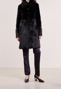 Sort faux fur frakke med krave, frontknapper og varierende teksturer; parret med stribede bukser og sorte højhælede sko.
