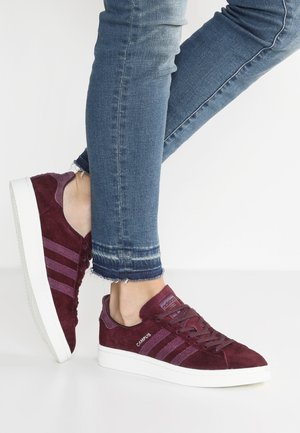 Osoba w dopasowanych niebieskich dżinsach i bordowych sneakersach Adidas Campus z białymi podeszwami, jedna stopa lekko uniesiona na jednolitym tle.