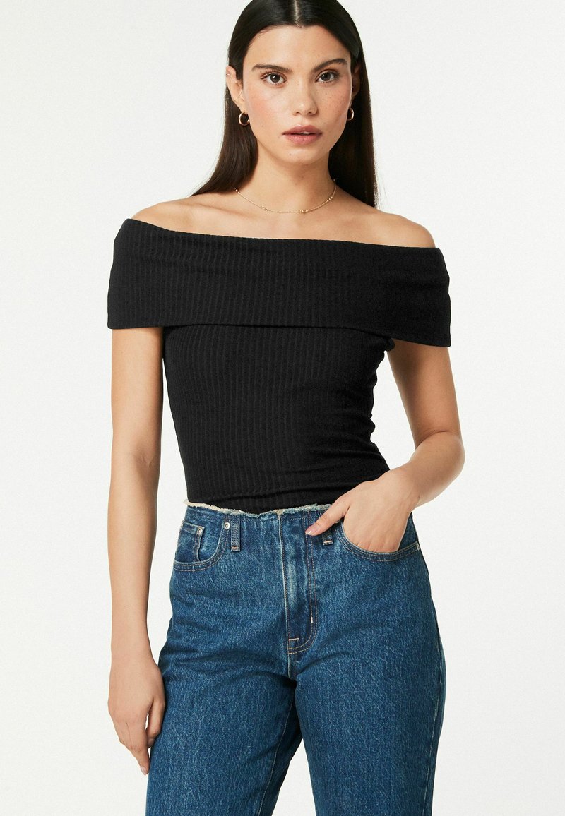 Melnā off-the-shoulder ribotā blūze, izgatavota no elastīga auduma, apvienota ar augsta jostasvietas zilu džinsu biksēm ar saplēstām malām.