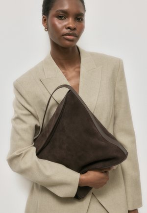 Beige värvi bleiserit kandev naine, kes hoiab suurt tumedat pruuni seemisnahast käekotti valgel taustal