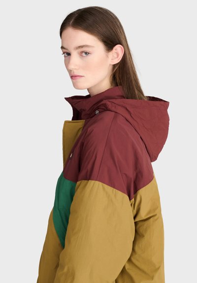 Jeune femme aux longs cheveux bruns portant une veste à capuche color block bordeaux, moutarde et vert, regardant vers la gauche sur un fond clair.