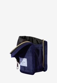 Marineblå velour kosmetikkveske med gullglidelås, som har en strukturert design og svart innvendig. Merket sier "THE FLAT LAY CO. LONDON."