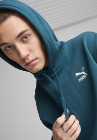 Felpa teal con coulisse e logo Puma bianco. Il tessuto sembra liscio, con una vestibilità comoda e cappuccio.
