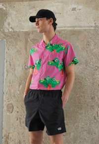 Camisa de botão cor-de-rosa de manga curta com padrões de rã verde; combinada com calções pretos com cintura elástica e bolsos laterais.