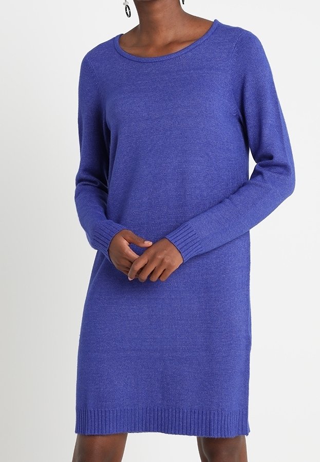 Donna che indossa un vestito a maglia viola con maniche lunghe, lunghezza al ginocchio, con polsini e orlo a costine, in piedi con le mani unite davanti.