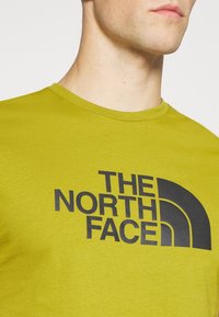 Groene T-shirt met korte mouwen van katoen, voorzien van een groot zwart logo en een gebogen ontwerpelement. Soepele textuur, ronde hals.
