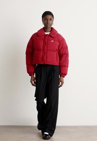 Κόκκινο κοντό πούφφερ μπουφάν με στρογγυλή γιακά, μπροστινά κουμπιά και καπιτονέ σχέδιο. Φορεμένο με μαύρα παντελόνια σε ευρεία γραμμή και μαύρα παπούτσια.