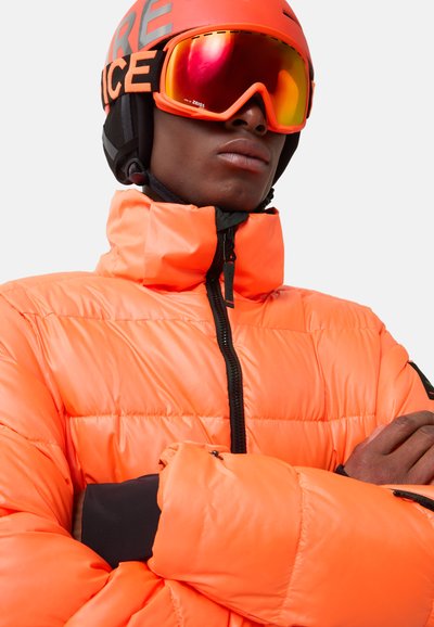 Pomarańczowa kurtka puchowa z wysokim kołnierzem, z czarnym zamkiem i mankietami. Czarne gogle narciarskie z lustrowanymi soczewkami i kask.