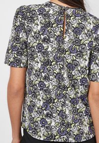 Korte mouwen blouse in wit, met een paars en groen bloemenpatroon, voorzien van een keyhole achterkant en textuurstof.