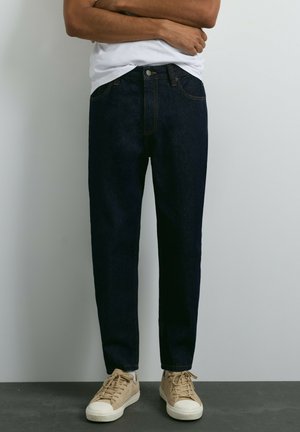 Homme portant un jean bleu foncé, un t-shirt blanc et des baskets beiges, debout les bras croisés devant un fond uni.
