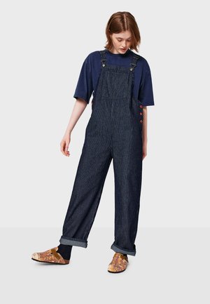 Mladá žena v navy modrém oversized tričku a tmavých pruhovaných overallových kalhotách s ohrnutými nohavicemi a barevnými slip-on botami, stojící na světlem pozadí.