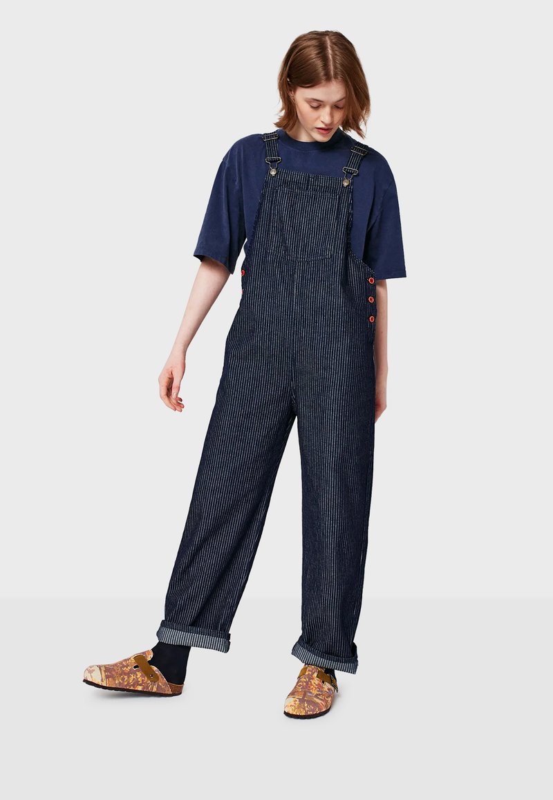 Young woman wearing navy blue oversized T-shirt and dark pinstriped overalls with cuffed legs and colorful slip-on shoes, standing on light background.