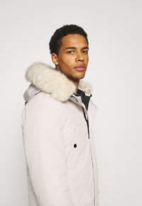 Giovane uomo che indossa un parka invernale beige chiaro con cappuccio bordato di pelliccia, che guarda verso la telecamera su uno sfondo bianco uniforme.