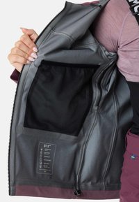 Vandtæt jakkeindvendig med gråt for, netlomme og vaskeanvisning. Yderskal i dyb bordeaux. Glat tekstur.