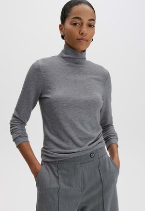 Femme portant un pull à col roulé gris et un pantalon gris, les mains dans les poches, debout devant un fond gris uni.
