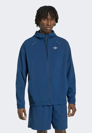 Homme avec de courtes dreadlocks portant une veste à capuche bleue zippée et un short assorti, regardant devant avec une expression neutre.