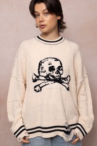 Joven con pelo corto y oscuro que lleva un suéter de punto color crema con un diseño de calavera y tibias cruzadas en negro y dobladillos desgastados.