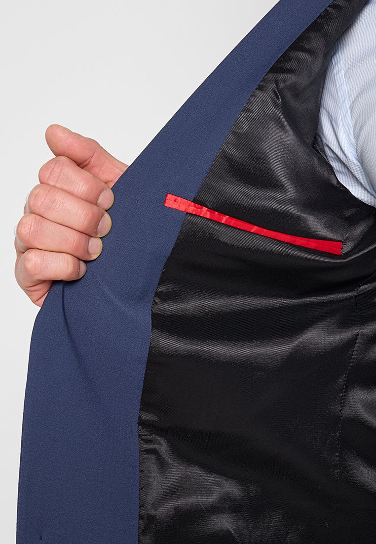 Interno della giacca da abito blu navy, con foderato in satin nero e dettagli rossi sottili sulle tasche. Una mano tiene il bordo della giacca.