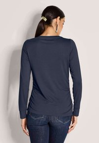 Vrouw met donker haar in een paardenstaart, gekleed in een marineblauwe longsleeve en donkerblauwe jeans, met de rug naar de camera gericht tegen een neutrale achtergrond.