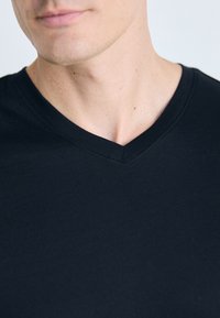 Gros plan sur le bas du visage et le cou d'une personne portant un t-shirt noir à col en V sur un fond clair.