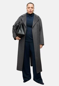 Cappotto lungo di lana grigia con collo a revers, indossato sopra un maglione blu navy e pantaloni a gamba larga. Tiene in mano una borsa di pelle nera. Gli accessori sono minimali.