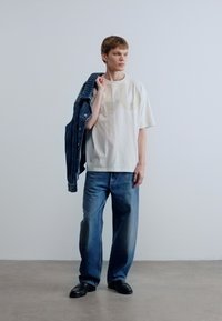 Άσπρο μεγάλο T-shirt με διακριτικό γραφικό, σκούρο τζιν μπουφάν κρεμασμένο στον ώμο, ανοιχτό μπλε άνετο τζιν, μαύρα παπούτσια.